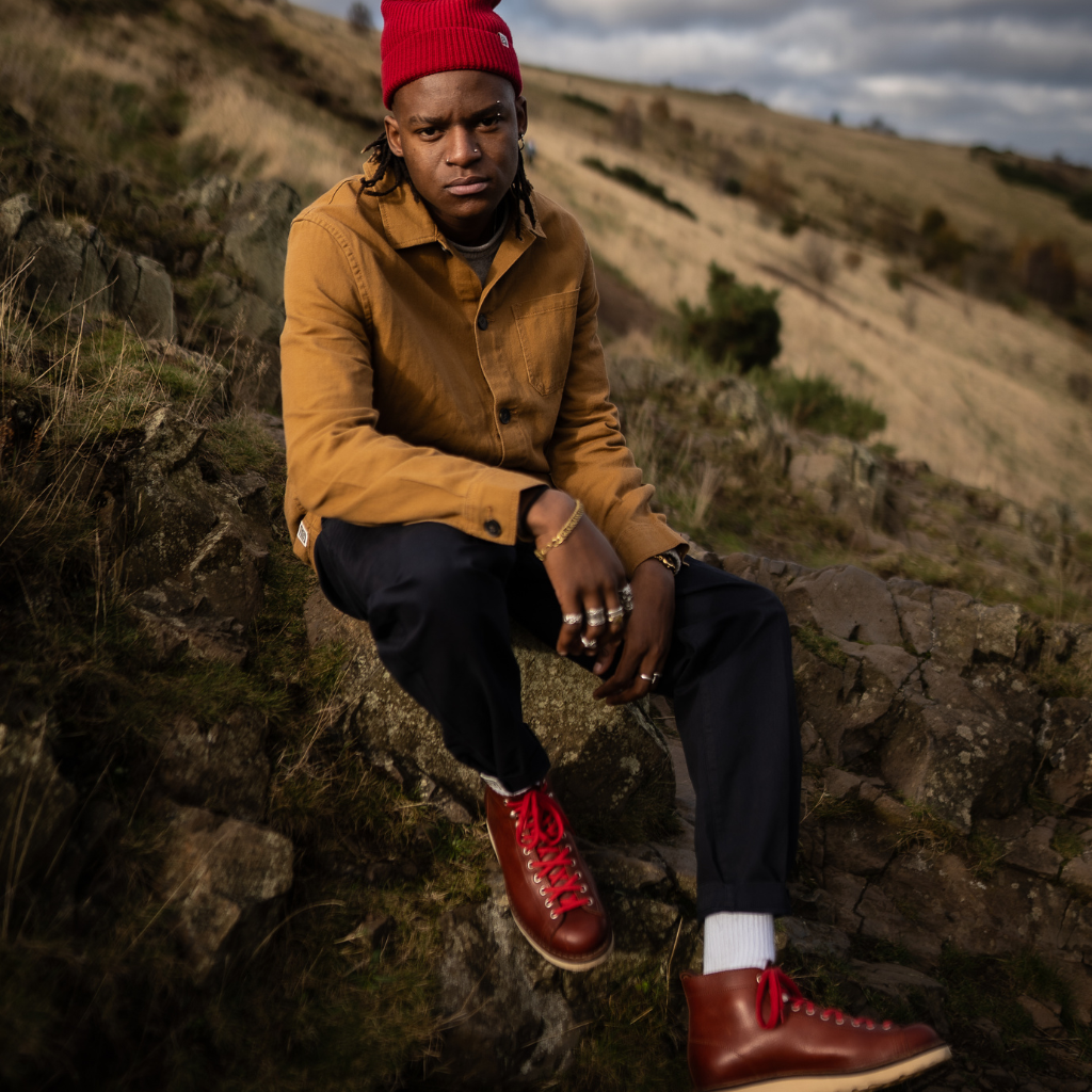 A person wearing a Paradaiza brown worker jacket with buttons, standing near a rock with greenery in the background.
