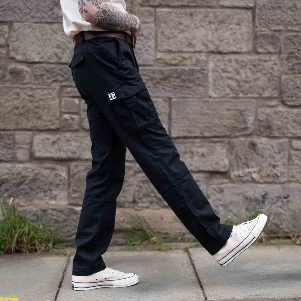 A person standing in an indoor setting wearing paradaiza black cargo pants and a grey shirt with sneakers.