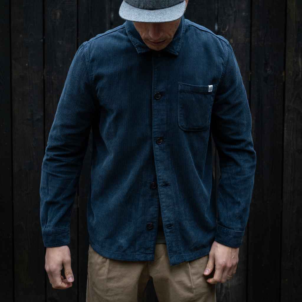 A male model wearing a paradaiza navy-colored corduroy overshirt with a collar, patch chest pocket, and buttons down the front, paired with beige pants.