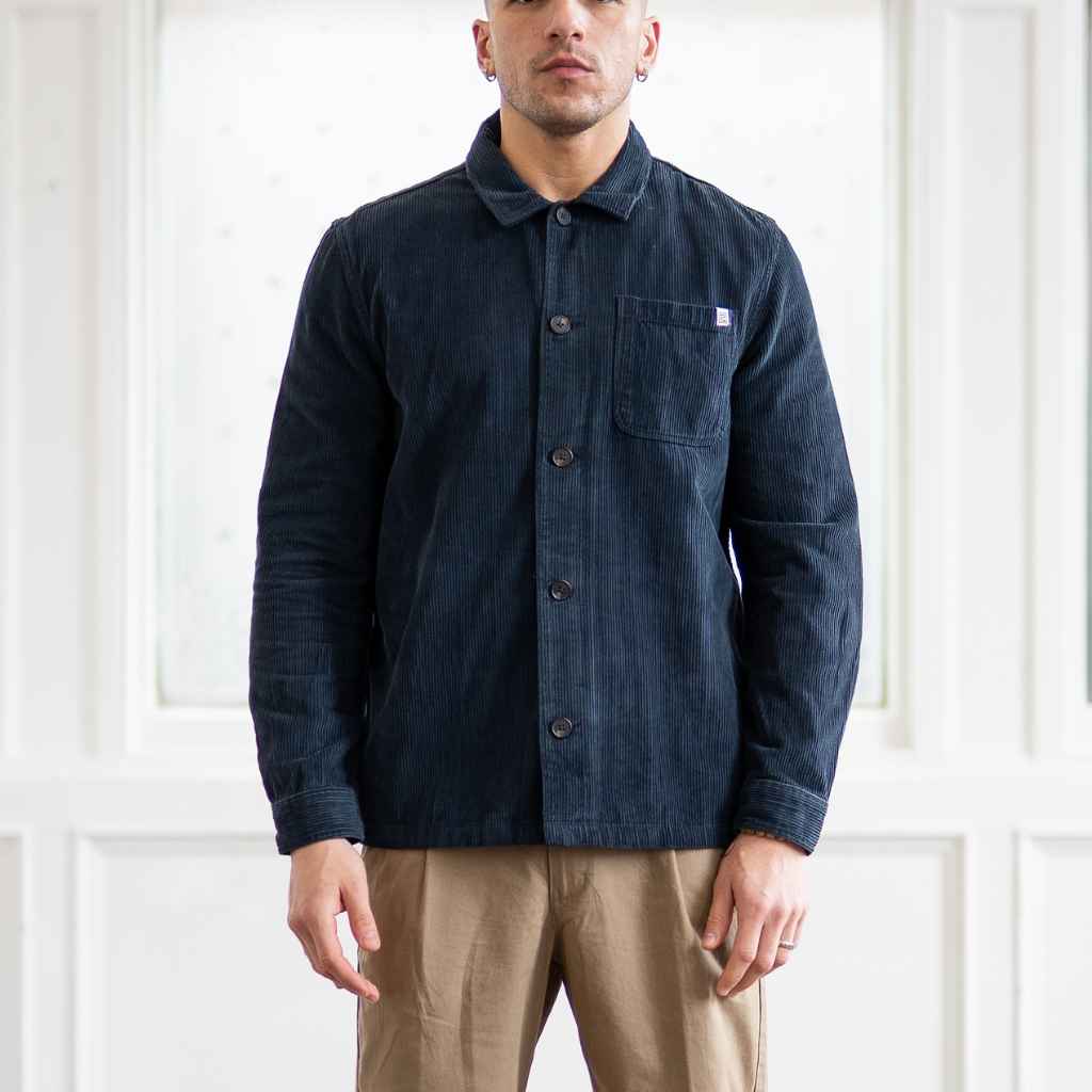 A male model wearing a paradaiza navy-colored corduroy overshirt with a collar, patch chest pocket, and buttons down the front, paired with beige pants.