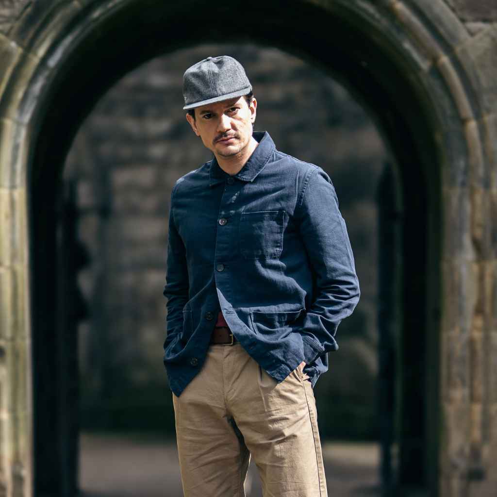 A man wearing a Paradaiza navy-colored worker jacket with long sleeves, featuring a collar, button-up front, and two patch pockets on the hem. Aswell as a paradaiza cap