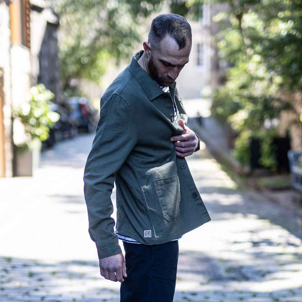 A man wearing an Paradaiza olive green worker jacket with buttoned front and collar, featuring two patch pockets and a round pleat on the back.