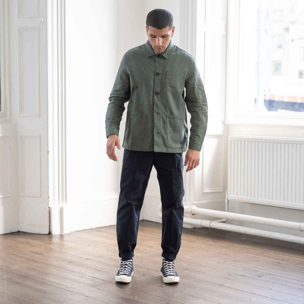 A man wearing an Paradaiza olive green worker jacket with buttoned front and collar, featuring two patch pockets and a round pleat on the back.