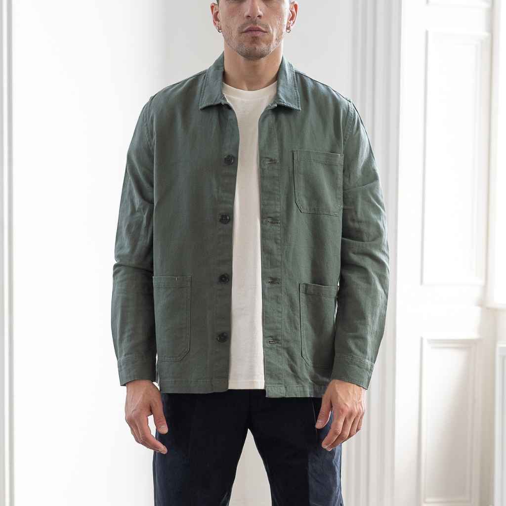 A man wearing an Paradaiza olive green worker jacket with buttoned front and collar, featuring two patch pockets and a round pleat on the back.