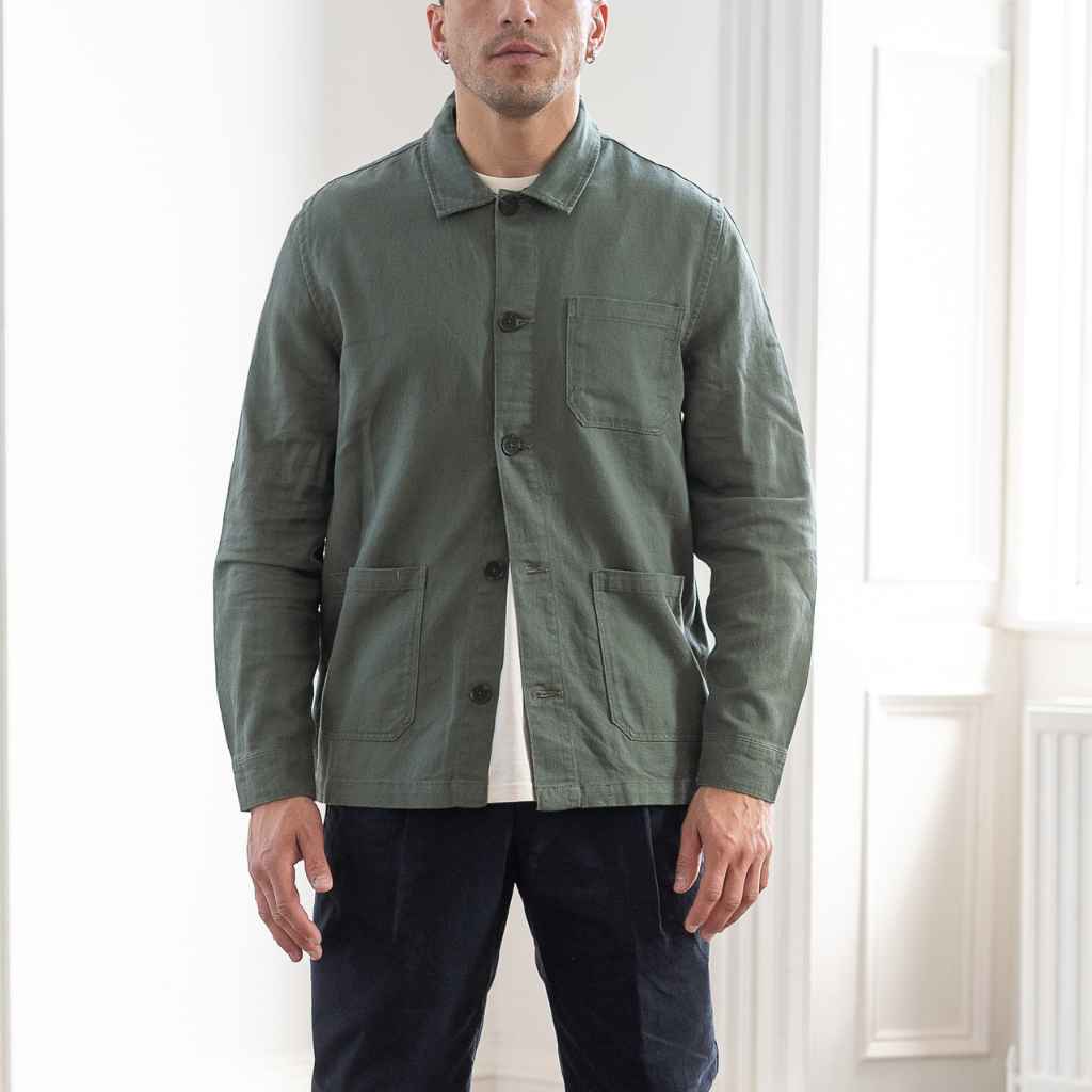 A man wearing an Paradaiza olive green worker jacket with buttoned front and collar, featuring two patch pockets and a round pleat on the back.