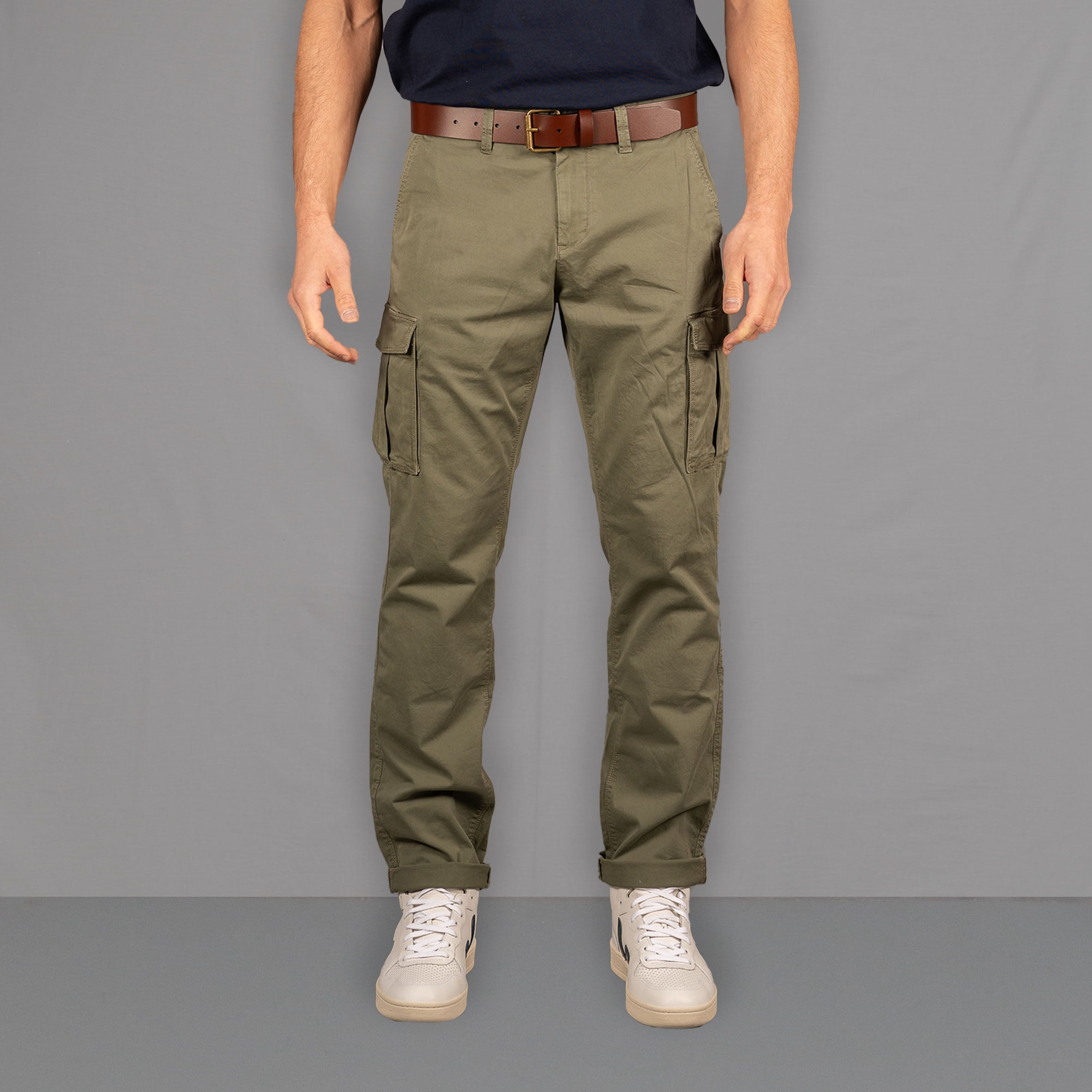 A person standing on a wooden floor wearing PARADAIZA olive green cargo pants with a belt loop, chain stitch detail, and multiple pockets, paired with a white shirt and white sneakers.