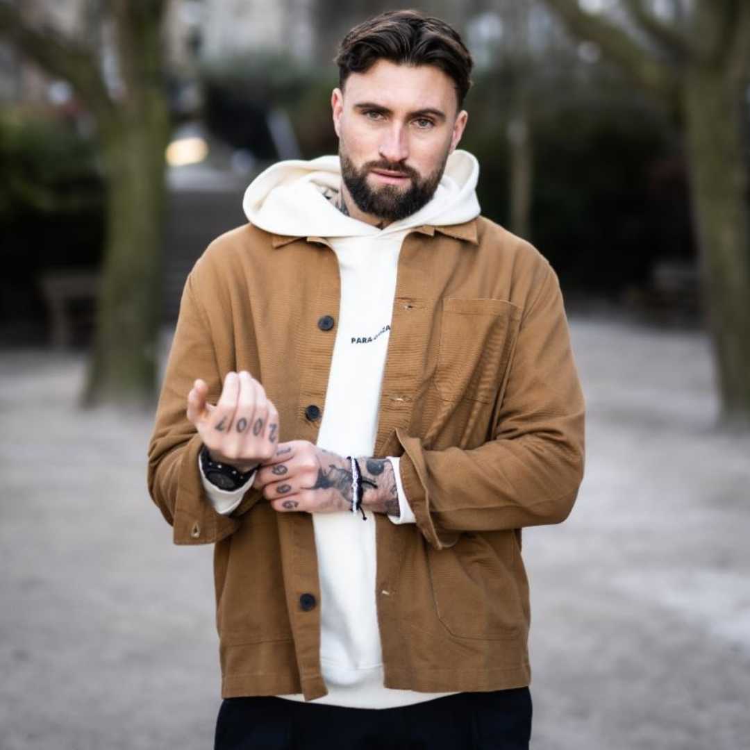 A person wearing a Paradaiza brown worker jacket with buttons and paradaiza hoodie, standing with greenery in the background.