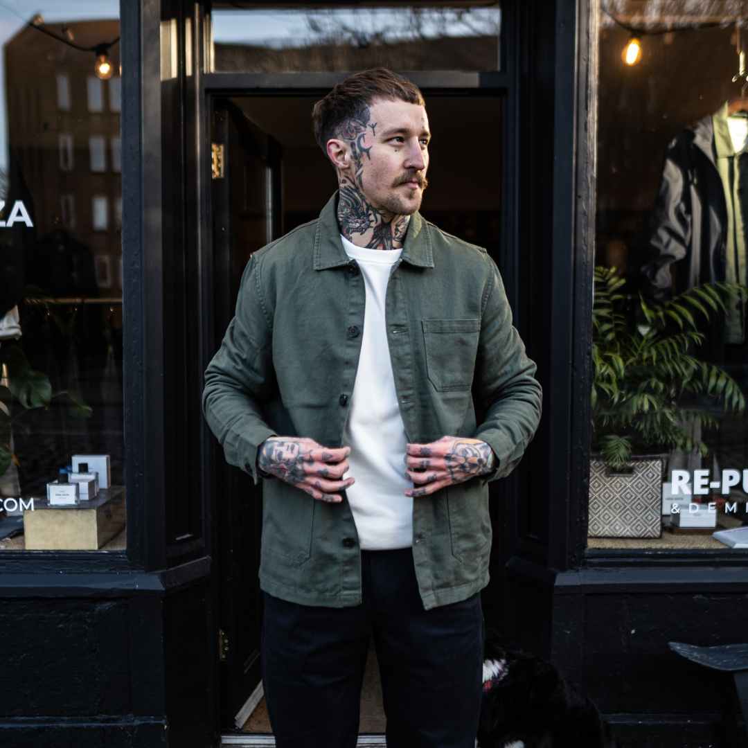 A man wearing an Paradaiza olive green worker jacket with buttoned front and collar, featuring two patch pockets and a round pleat on the back.