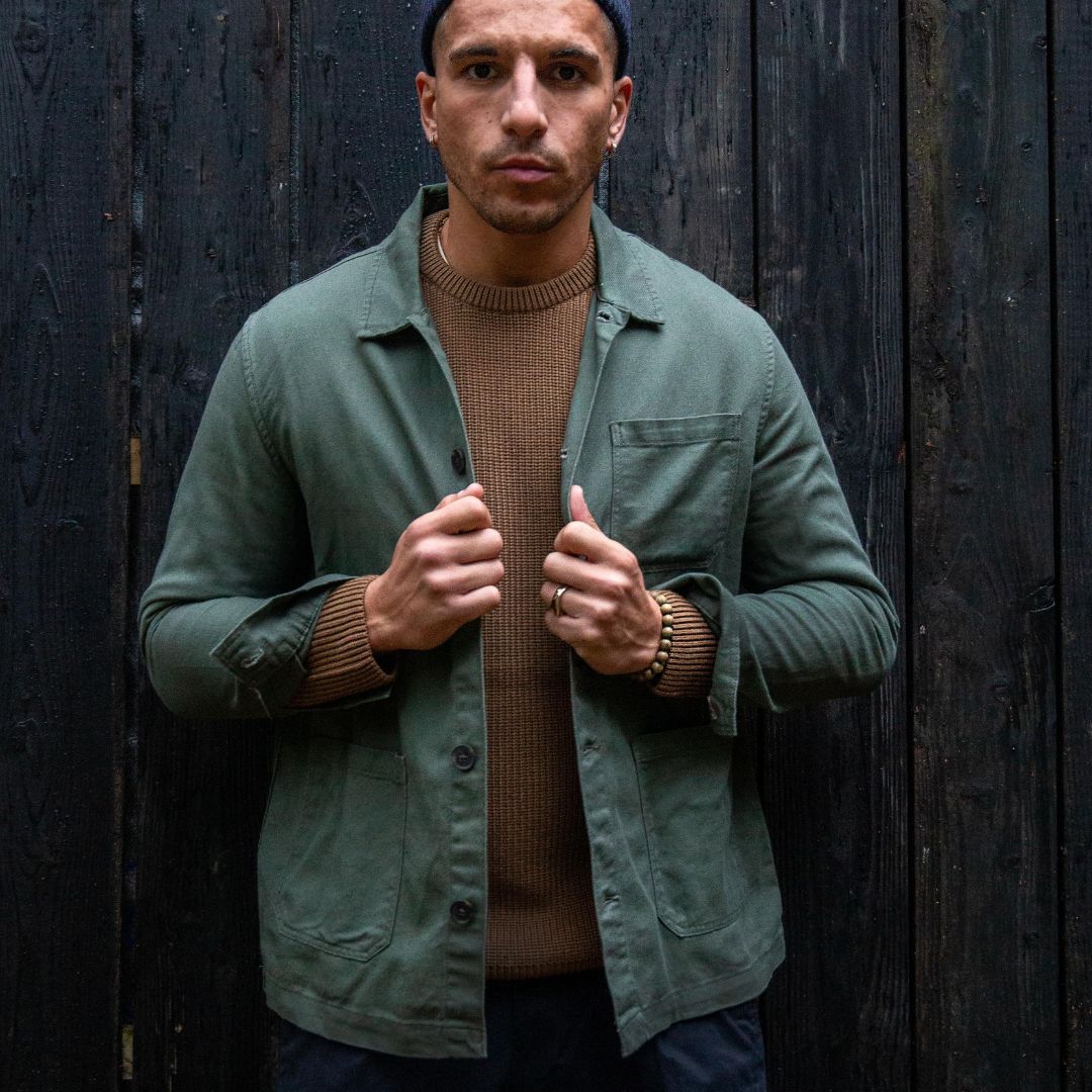 A man wearing an Paradaiza olive green worker jacket with buttoned front and collar, featuring two patch pockets and a round pleat on the back.