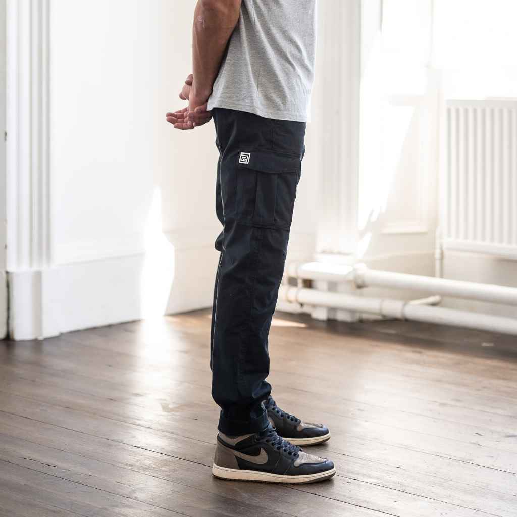 A person standing in an indoor setting wearing paradaiza black cargo pants and a grey shirt with sneakers.