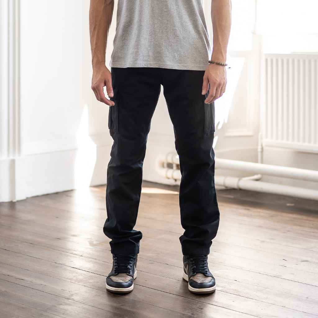 A person standing in an indoor setting wearing paradaiza black cargo pants and a grey shirt with sneakers.