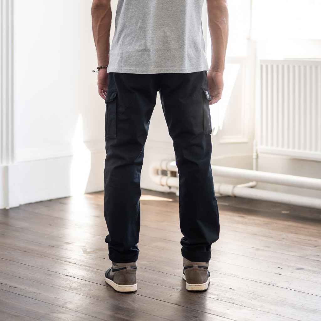 A person standing in an indoor setting wearing paradaiza black cargo pants and a grey shirt with sneakers.