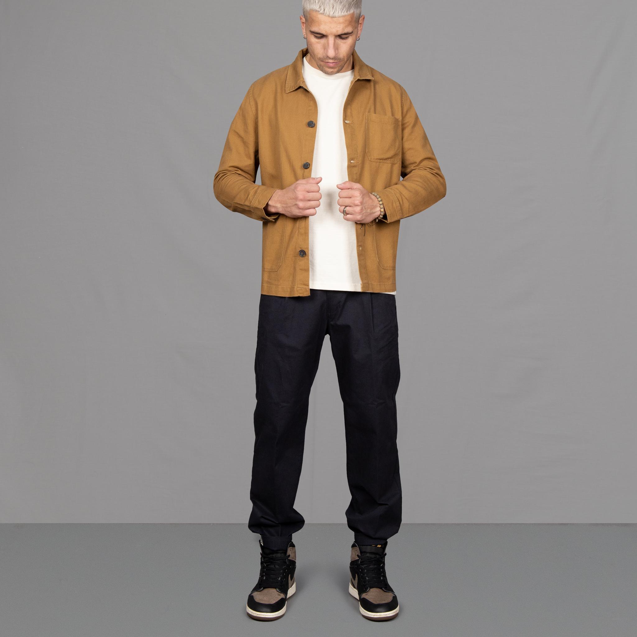 Model wears Para-Daiza Brown worker jacket , off-white tee, pinch chino and Nike trainers on grey floor.