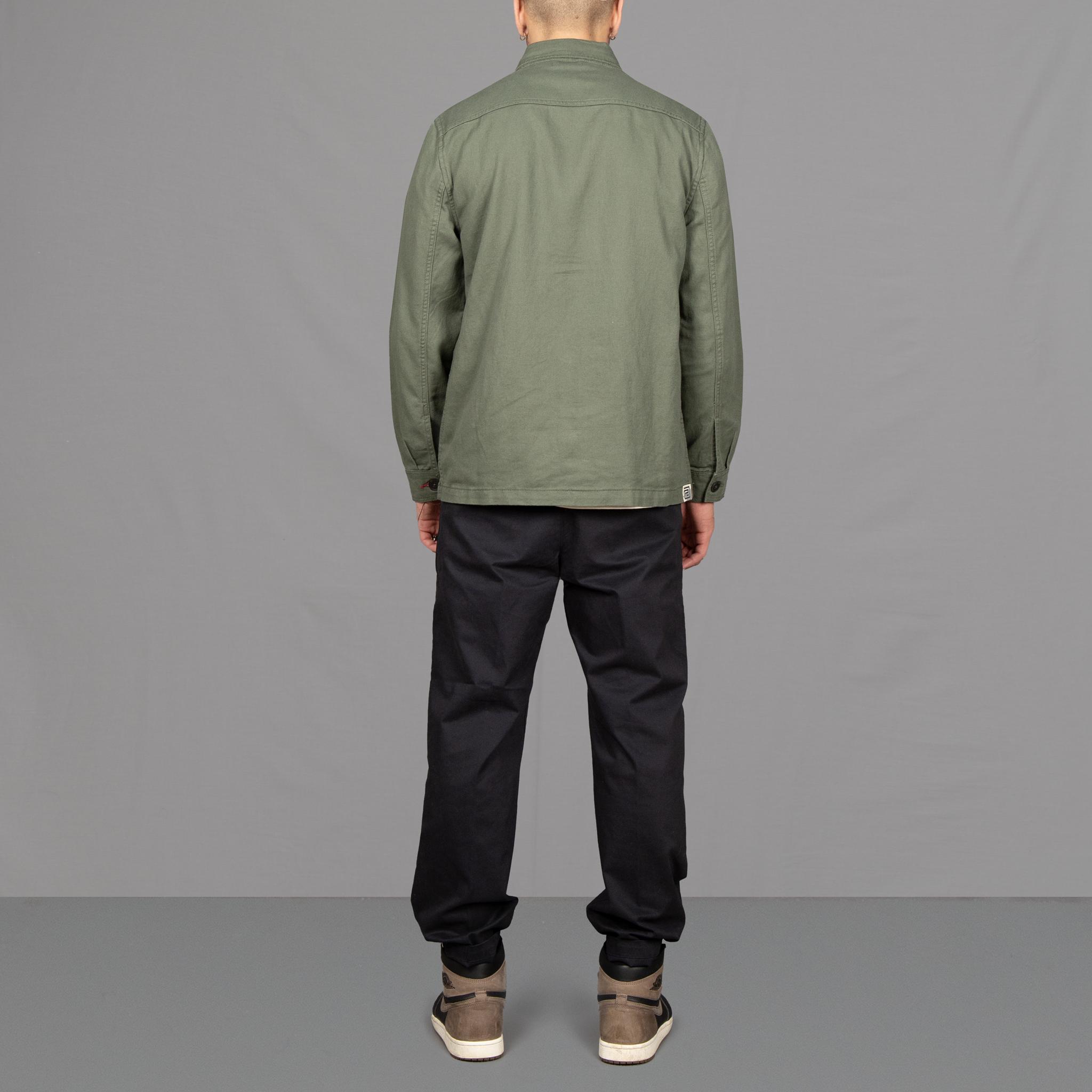 A man wearing an Paradaiza olive green worker jacket with buttoned front and collar, featuring two patch pockets and a round pleat on the back.