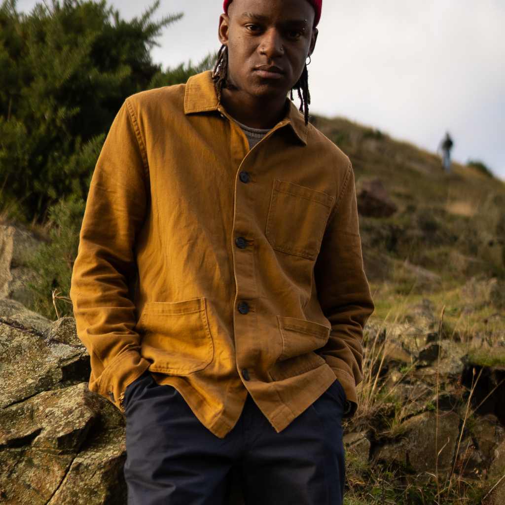 A person wearing a Paradaiza brown worker jacket with buttons, standing near a rock with greenery in the background.