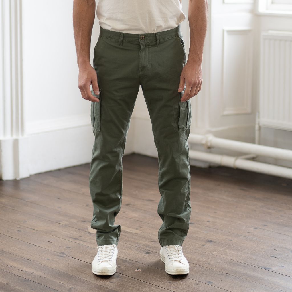 A person standing on a wooden floor wearing PARADAIZA olive green cargo pants with a belt loop, chain stitch detail, and multiple pockets, paired with a white shirt and white sneakers.