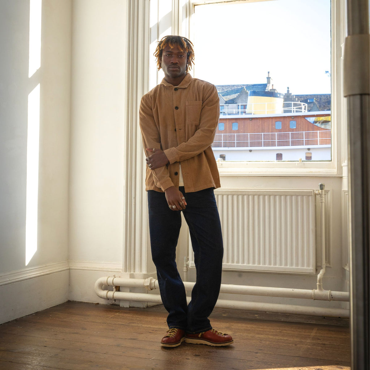 A man stands in a bright room with a large window, wearing a tan corduroy button-up shirt, dark navy trousers, and brown leather shoes.