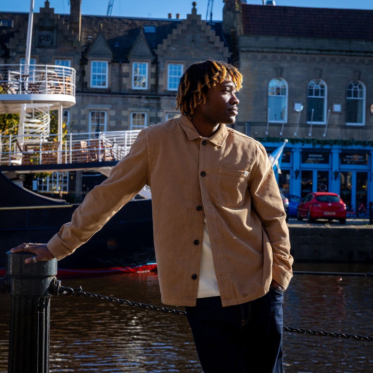 A man wearing a tan corduroy overshirt over a white t-shirt and dark jeans, standing by a canal with historic stone buildings and a boat in the background.
