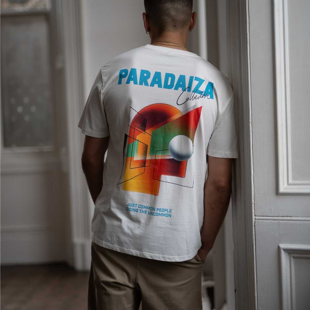 Person wearing a paradaiza white t-shirt with colorful design and text, standing against a brick wall.