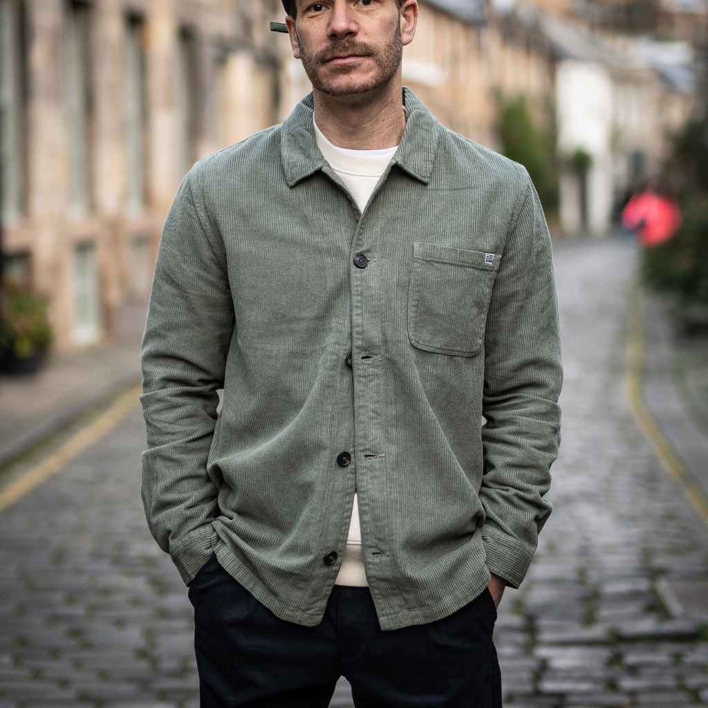A person wearing a paradaiza khaki-colored corduroy overshirt with a button-up collar, a patch chest pocket, and holding a matching green cap.