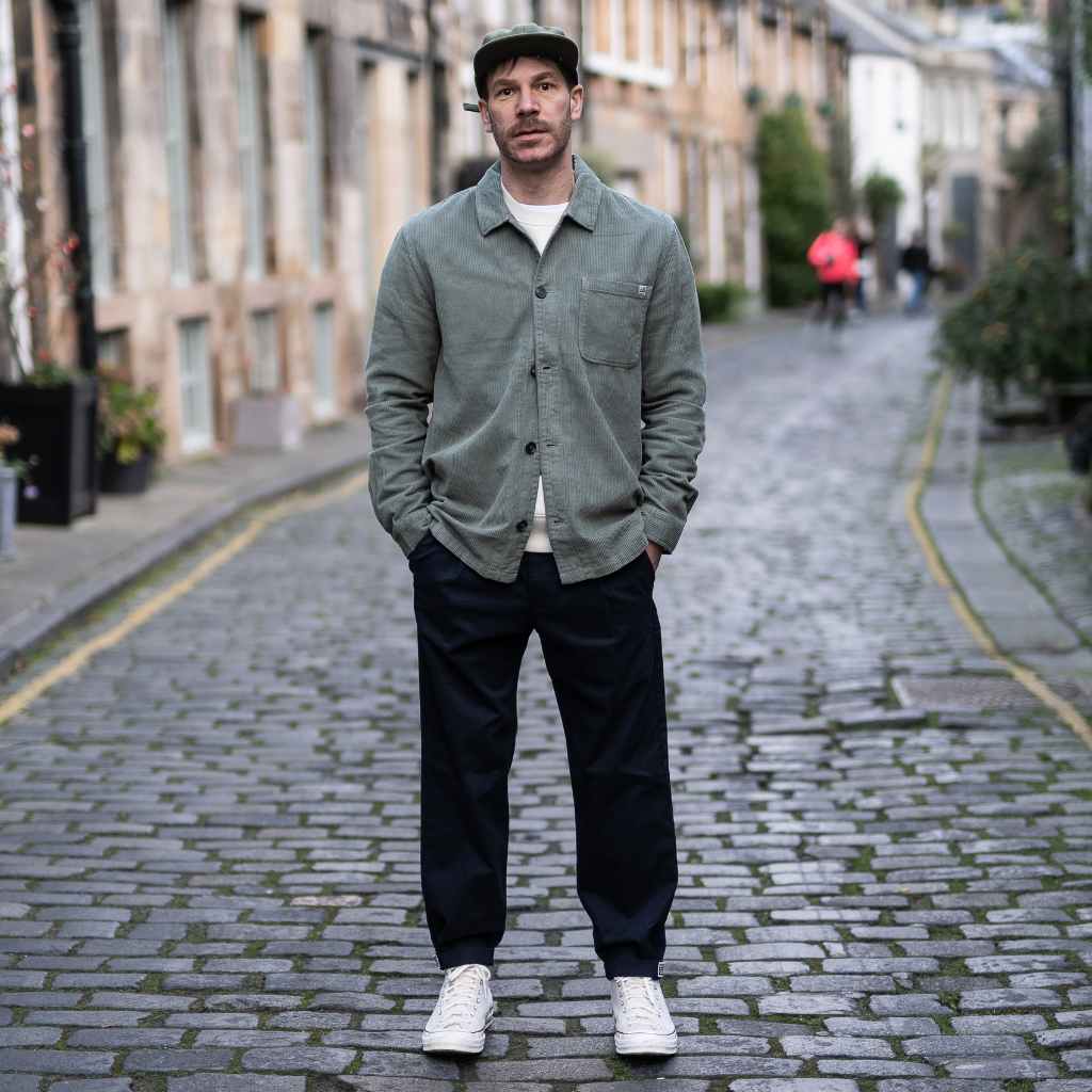 A person wearing a paradaiza khaki-colored corduroy overshirt with a button-up collar, a patch chest pocket, and a matching green cap.