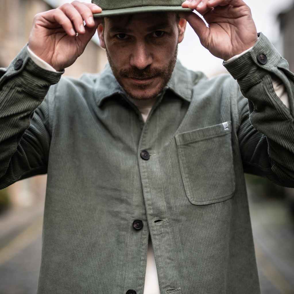 A person wearing a paradaiza khaki-colored corduroy overshirt with a button-up collar, a patch chest pocket, and holding a matching green cap.