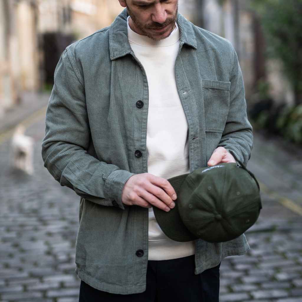 A person wearing a paradaiza khaki colored corduroy overshirt with a button-up collar, a patch chest pocket, and holding a matching green cap.