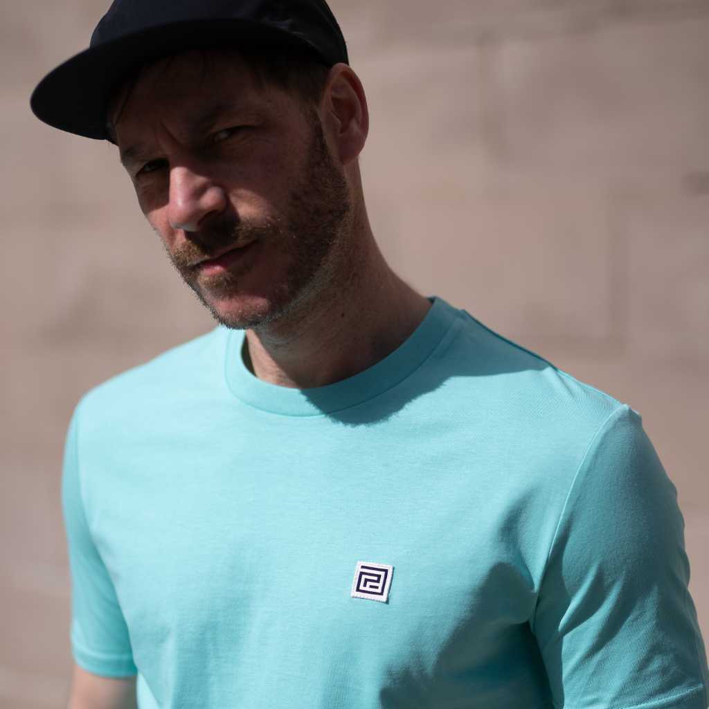 Man wearing a paradaiza ocean blue t-shirt with a small logo against a wall.