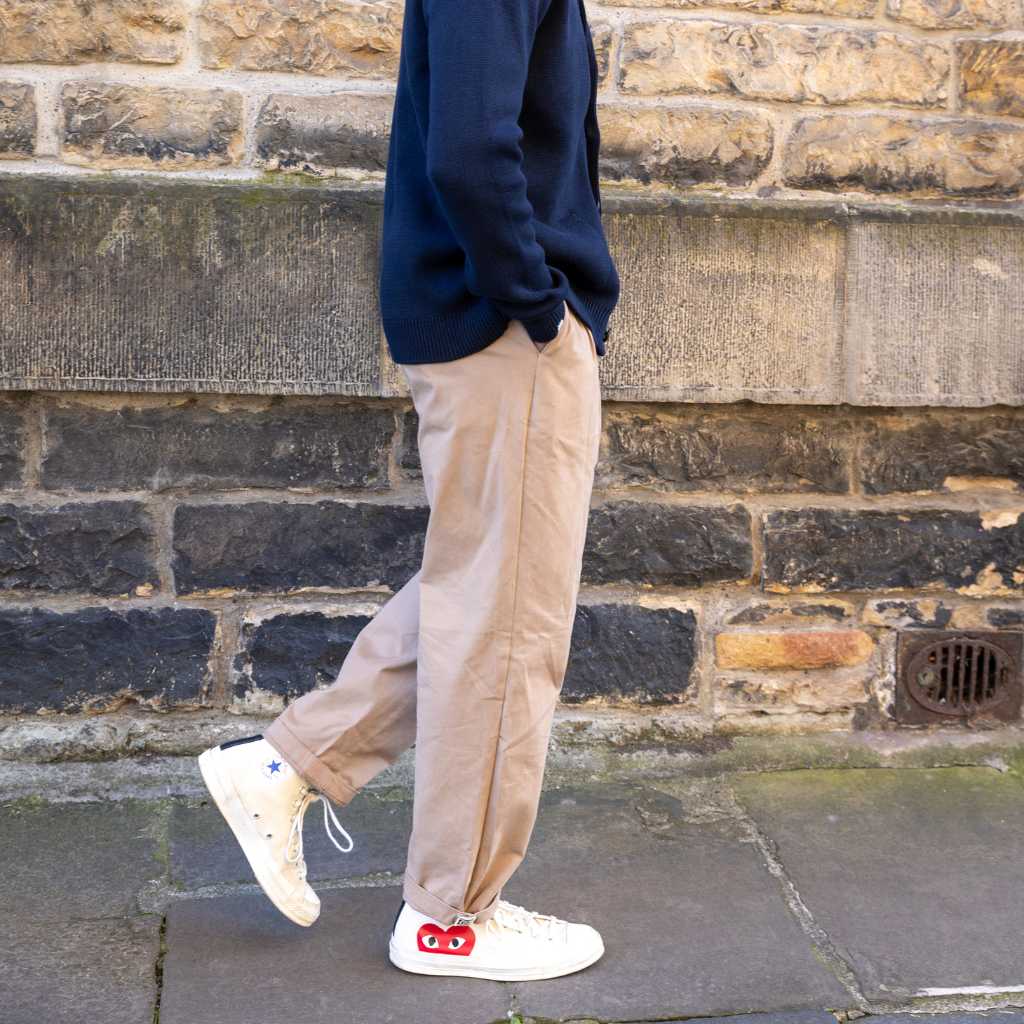 Person wearing beige paradazia pinch chino, and white sneakers with red accents against a brick wall.