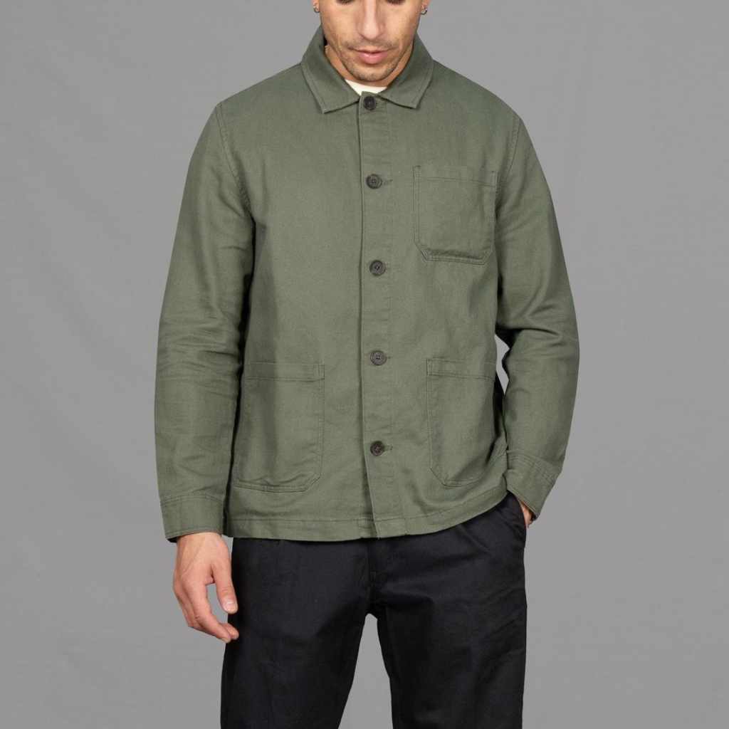 A man wearing an Paradaiza olive green worker jacket with buttoned front and collar, featuring two patch pockets and a round pleat on the back.