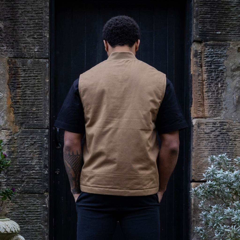 Man wearing a paradaiza tan vest over a black shirt standing in front of a dark door.