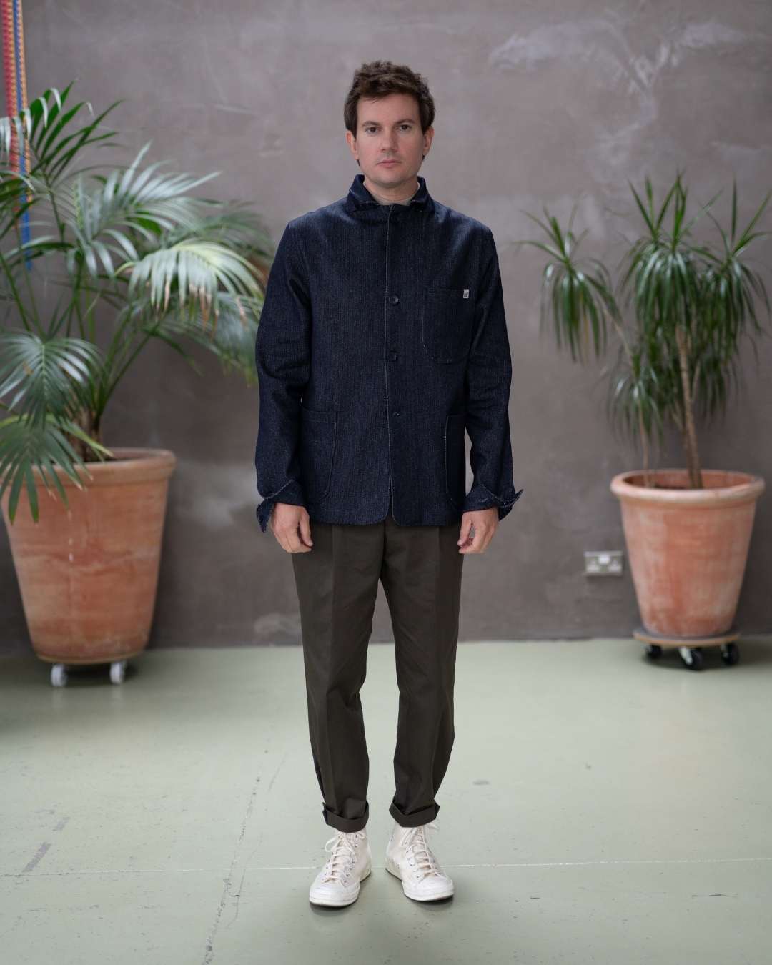 Model  wearing a dark denim paradaiza  jacket and khaki pinch chinos, standing indoors with plants in the background.
