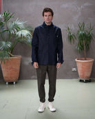 Model  wearing a dark denim paradaiza  jacket and khaki pinch chinos, standing indoors with plants in the background.