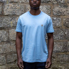 Person wearing a sky blue paradaiza t-shirt with a small logo against a stone wall.