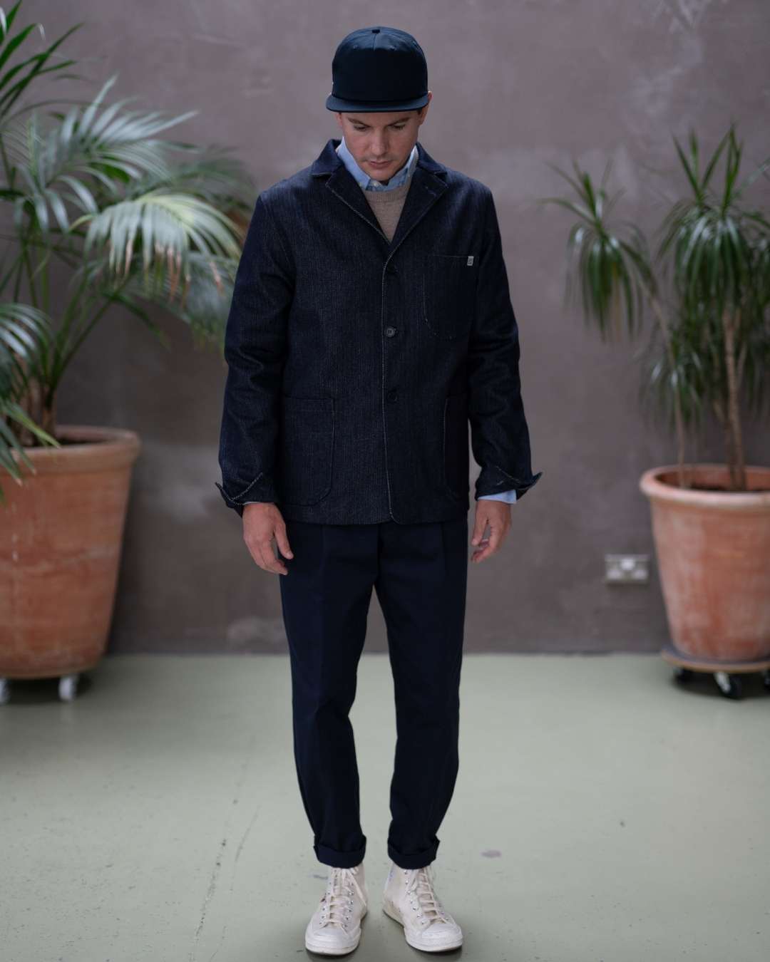 Model  wearing a dark denim paradaiza  jacket and pants with a cap, standing indoors with plants in the background.