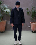Model  wearing a dark denim paradaiza  jacket and pants with a cap, standing indoors with plants in the background.