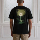 Person wearing a black paradaiza  t-shirt with a forest design on the back, standing against a white paneled wall.