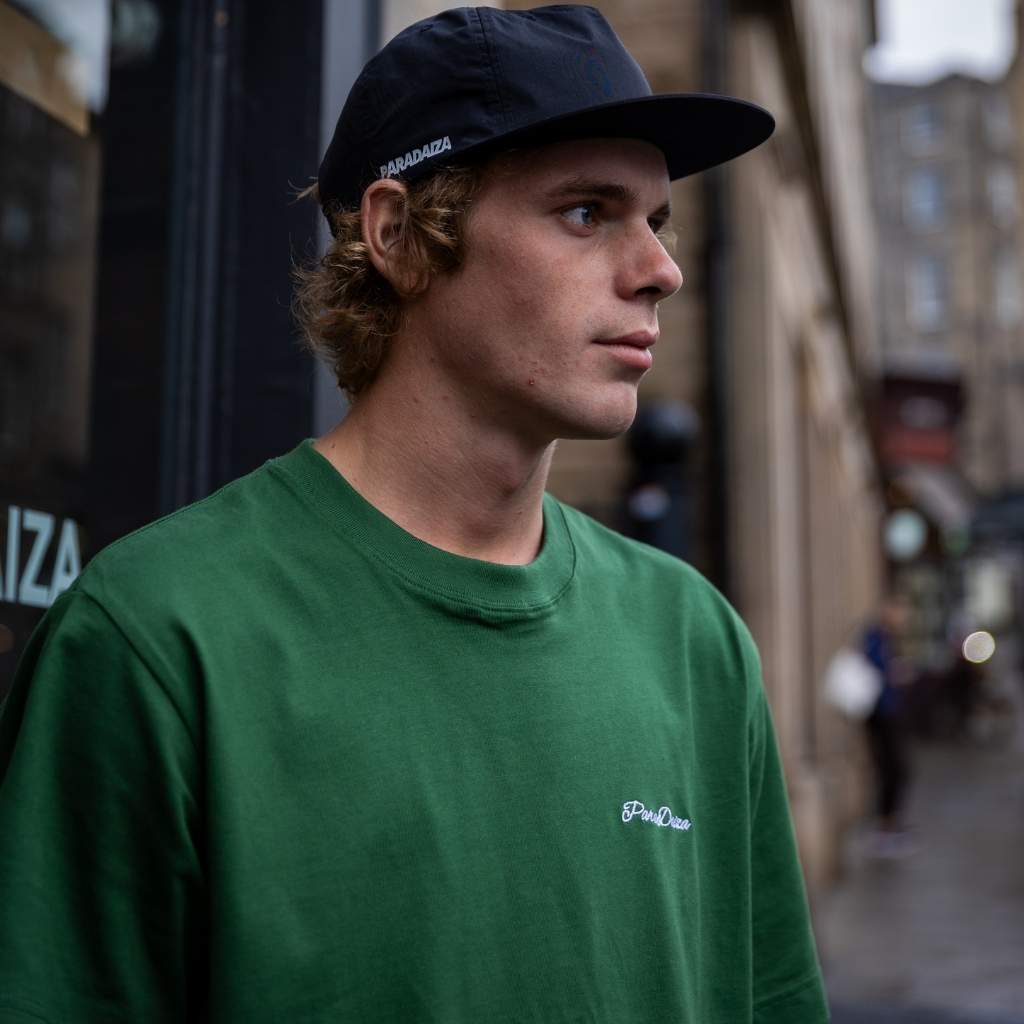 A man wearing a dark green crewneck sweatshirt with a small white embroidered logo on the left chest and a black baseball cap, standing on a blurred city street.
