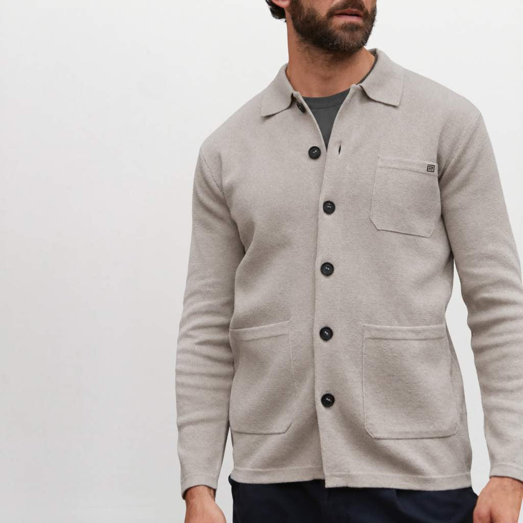 A man wearing a beige Paradaiza  knit button-up shirt jacket with a collar, black buttons, a chest pocket, and two large front patch pockets, paired with a dark gray t-shirt and navy pants, against a plain light gray background.