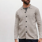 A man wearing a beige Paradaiza  knit button-up shirt jacket with a collar, black buttons, a chest pocket, and two large front patch pockets, paired with a dark gray t-shirt and navy pants, against a plain light gray background.
