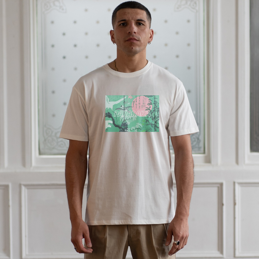 Man wearing a paradaiza white t-shirt with a graphic design in front of a white paneled wall.