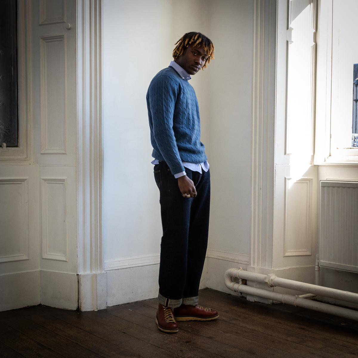 A man stands in a sunlit room with white paneled walls and dark wood floors, wearing a blue cable-knit sweater over a light blue collared shirt, dark trousers, and brown leather shoes.