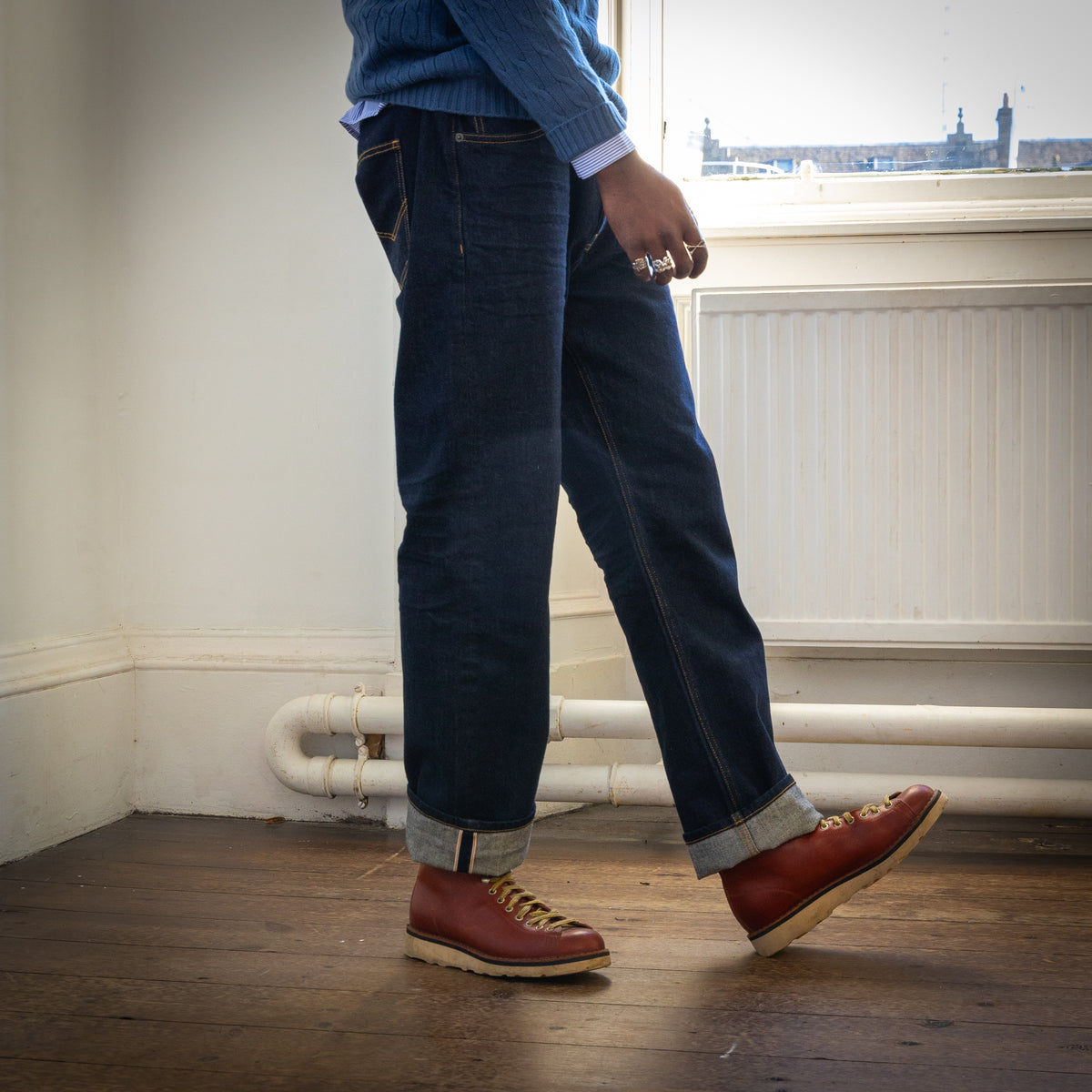 A person wearing dark blue denim jeans with rolled cuffs, a blue knit sweater, and brown leather lace-up boots, standing on a wooden floor in front of a white wall and window.