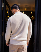 Man wearing a  paradaiza beige sweater standing in front of a dark doorway with lights.