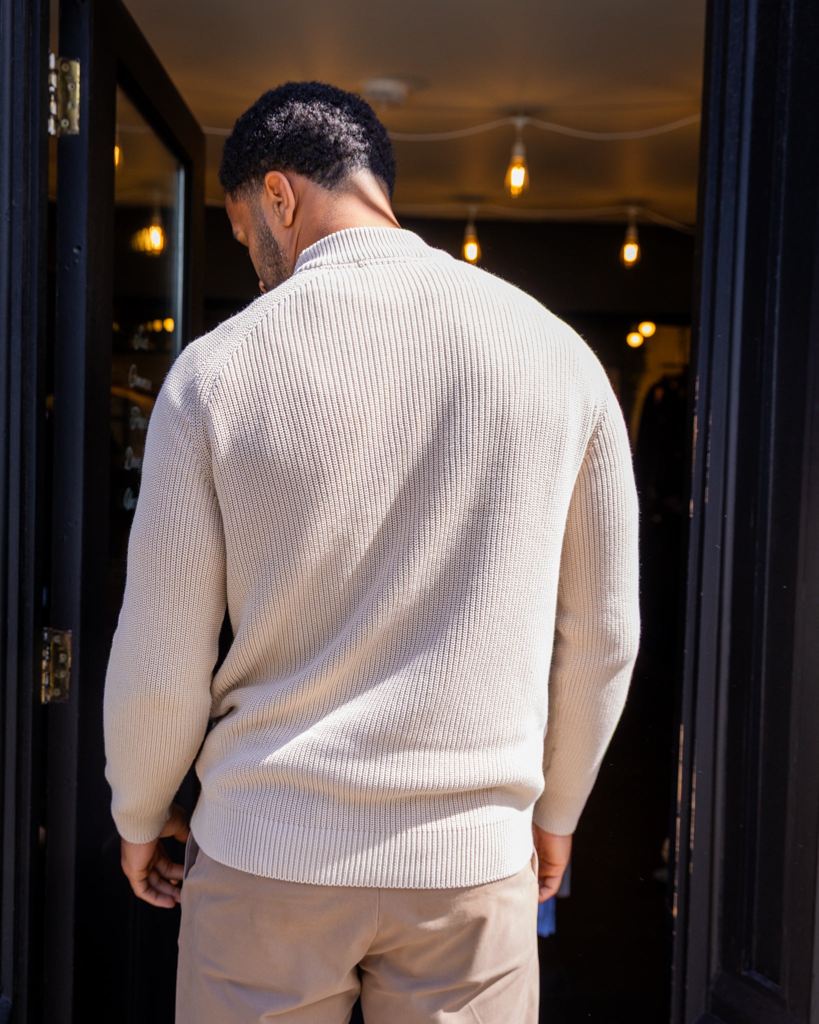 Man wearing a  paradaiza beige sweater standing in front of a dark doorway with lights.