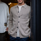 A man wearing a light beige wool-blend button-up cardigan over a white t-shirt and navy pants, standing in a doorway. Another person in a white shirt and grey cap is partially visible on the left.