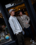 Two men smiling and posing in a storefront doorway. The man on the left wears a white long-sleeve button-up shirt and a light cap, while the man on the right wears a beige button-up shirt over a white tee with dark pants.