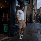 A man stands on a city sidewalk wearing a white crew-neck t-shirt, beige drawstring shorts, purple socks, and white high-top sneakers with red laces. He is posed in front of a dark storefront on a wet street.