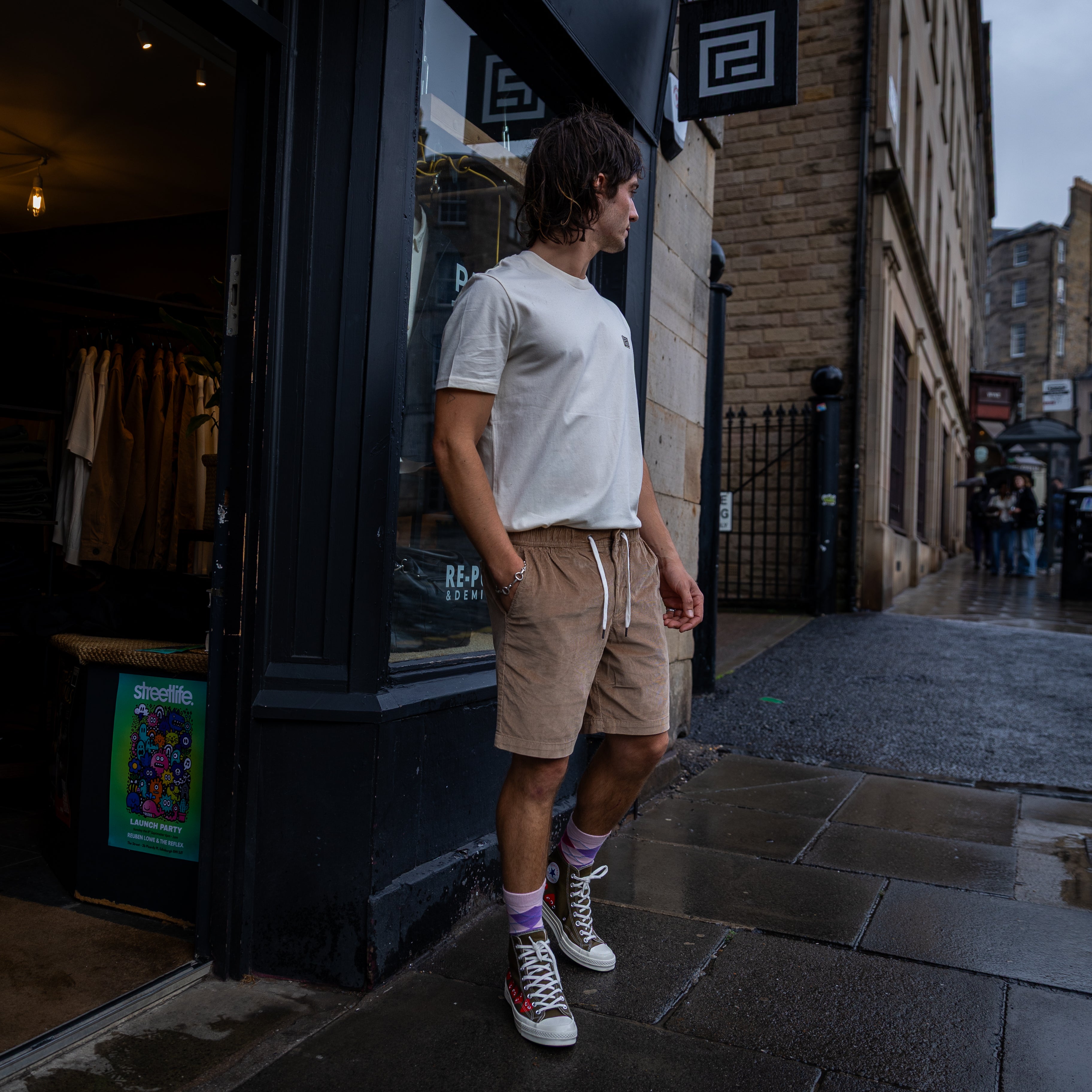 A man stands on a city sidewalk wearing a white crew-neck t-shirt, beige drawstring shorts, purple socks, and white high-top sneakers with red laces. He is posed in front of a dark storefront on a wet street.