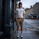 A man stands on a city sidewalk wearing a white crew-neck t-shirt with a small chest logo, tan drawstring shorts, purple socks, and white high-top sneakers. The background shows a wet street with brick buildings and a double-decker bus.